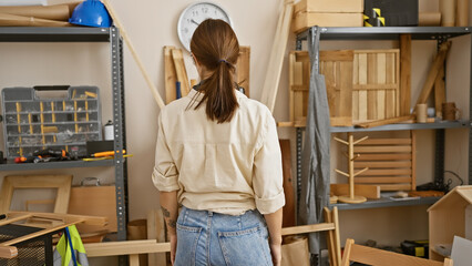 Back view of a young woman standing thoughtfully in a well-equipped carpentry workshop.