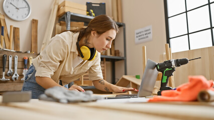 Focused woman using laptop in a carpentry workshop with tools and wood, embodying a capable artisan...