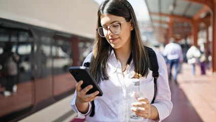 Young beautiful hispanic woman walking down the railway using smartphone at train station - obrazy, fototapety, plakaty