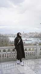 A smiling woman in a black coat poses with istanbul's skyline in the background, capturing travel and cultural exploration.