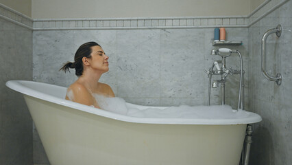 A relaxed woman enjoying a bubble bath in a clean, homely bathroom with bright lighting.