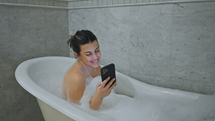 Smiling woman enjoying a bubble bath while using smartphone in a modern bathroom.