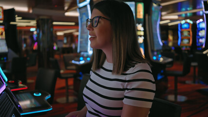 A smiling young woman in glasses plays slot machines in the vibrant ambiance of a casino.