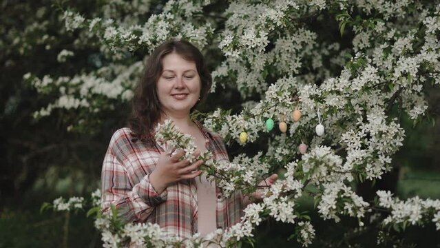 Inspired woman near flowering tree enjoys aroma and spring mood. Portrait of woman admiring white flowers and smiles at nature. Multi-colored small colored eggs decorate the branches.