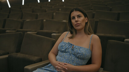 A young adult woman sits in a dimly lit theater, exuding elegance and anticipation in her blue...