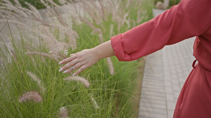 A young woman in a red dress gently touches tall grass while walking in a lush park.