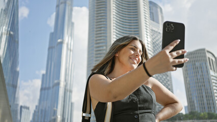 A smiling woman takes a selfie with a smartphone against the backdrop of the tall dubai skyscrapers, showcasing urban tourism.