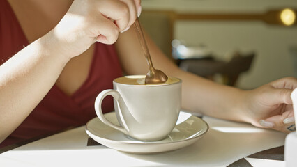 A young adult woman stirs coffee in a bright cafe, showcasing a casual indoor dining experience.