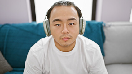Handsome young chinese man with a serious face engrossed in his world of music while relaxing at home, immersed in the serene ambiance of his indoor apartment, lost in the audio sound of his gadget.