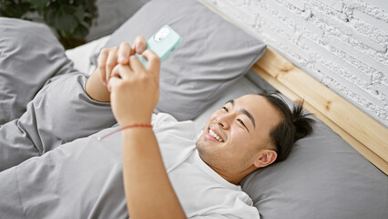 Cheery young chinese man in pyjamas luxuriating in a cozy bedroom, using smartphone for fun morning texting, confident asian adult, with pigtail hairstyle, chilling on comfortable bed.