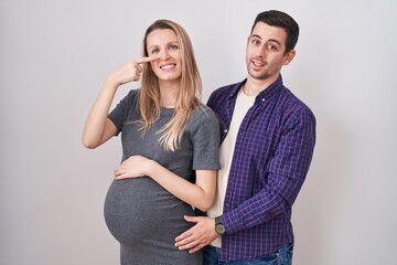 Young couple expecting a baby standing over white background pointing with hand finger to face and nose, smiling cheerful. beauty concept