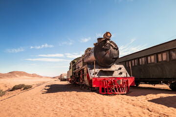 an old black, green and red vintage steam train rests on the tracks in a wild and sunny desert environment, with carriages in the background