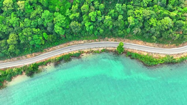 An epic drone shot captures a tropical coastal road, flanked by vibrant greenery and crystal-clear waters. Transportation and vehicles concept. Nature stock footage. Ko Chang island, Thailand. 4K.
