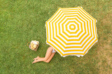 Woman in white bikini with basket for picnic lying under yellow umbrella on the green grass...
