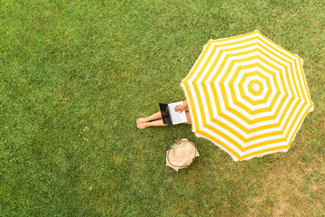 Woman with laptop on the knees sitting on the green grass under yellow umbrella sunbathes at summer day. Top view, drone, aerial view. - 721525686