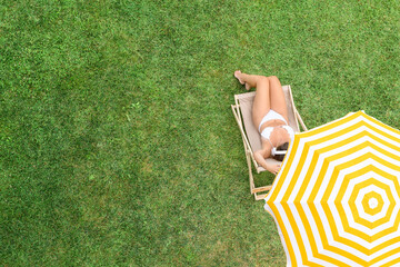 Woman in a white bikini sitting on deck chair under yellow umbrella  on the green grass sunbathes...