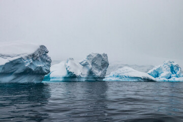 Rugged coastline covered in blue ice, on the Antarctic Peninsula. Gray overcast sky; snow falling. Calm, dark blue water in foreground. 
