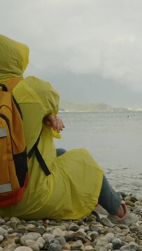 A woman in a yellow raincoat with a backpack sits and throws a stone into the sea. Rainy weather and overcast sky. Loneliness and sadness.