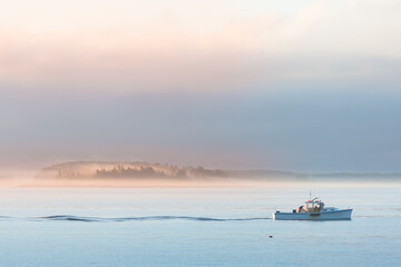 Lobster boat setting traps at sunrise along the coast of Maine. Fog covered islands in distance reflecting rising sun.