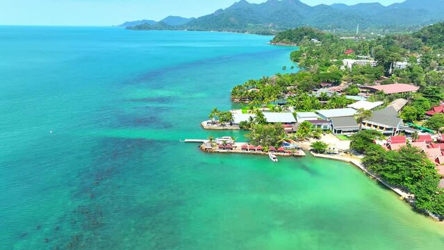 Breathtaking coastal beauty with sandy beaches, turquoise sea, resort-lined shores, lush mountain backdrop, and a serene blue sky. Shot from a drone's perspective. Mu Ko Chang National Park, Thailand.