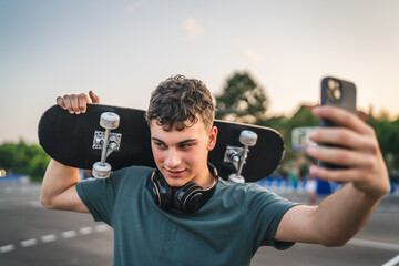 One man young adult caucasian teenager stand outdoor with skateboard on his shoulder and headphones...