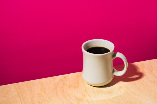 A white diner style coffee mug filled with black coffee on the edge of a wooden table against a red backdrop