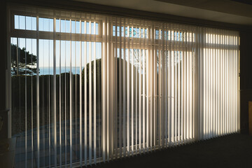 Sunlight shining through full length white vertical blinds in front of three glass sliding french doors leading to a patio, garden and sea view - 721592832