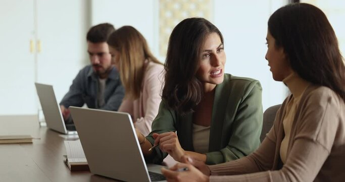 Woman colleagues discuss business project idea use laptop, sit at desk in coworking space, brainstorm, learn new business application, cooperate. Mentor explain employee online strategy on computer
