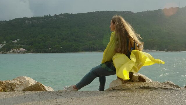 Strong wind and overcast weather in the sea bay. A woman in a yellow raincoat with a backpack sits on a rock in the bright sunlight.