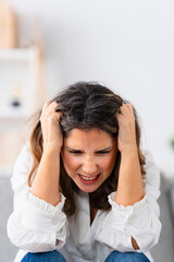 Furious woman sitting on the sofa at home hands on head and screaming