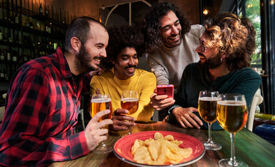 Multiethnic group of four male friends gathering in a bar drinking beer and looking at one phone together watching social media and having fun.