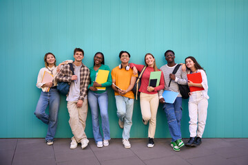 Cheerful international students posing smiling standing looking at the camera. Young people in multicultural community of friends and embracing with backpacks and workbooks in a blue wall background