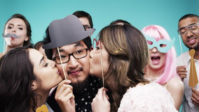 Happy people, party and kiss man on cheek, moustache and event celebration with excited friends. Dancing, holiday and photobooth with men, woman and social gathering on studio background for fun