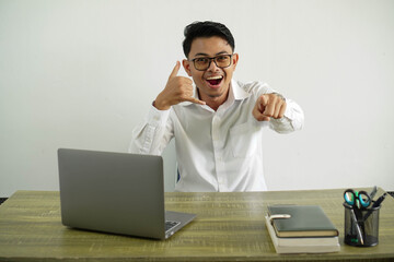 young asian businessman in a workplace making phone gesture and pointing front, wearing white shirt with glasses isolated