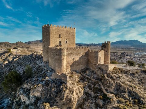 Aerial view of Jumilla medieval castle in Murcia Spain, on a hilltop, imposing irregular shape keep with four floors, crenelated battlements cloudy blue sky background
