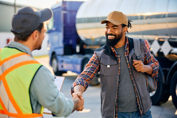 Happy black truck driver and freight transportation manager handshaking on parking lot.