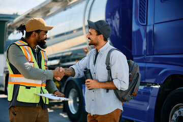 Happy truck driver and black freight transportation manager handshaking on parking lot.