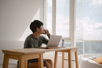 Asian man working with computer in apartment with city view at the windows in morning light.