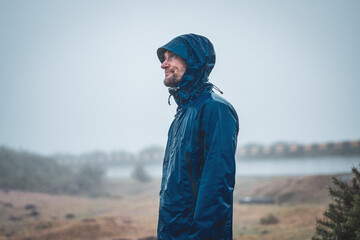 Disillusioned and healthy male tourist with hands in trouser pockets looking at rainy sky with...