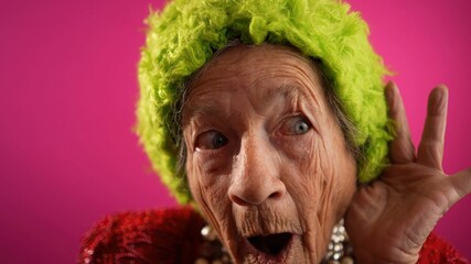 Funny fisheye view of smiling happy crazy grandmother with no teeth and wrinkled skin puts hand to ear to listen to a secret in isolated on pink background studio