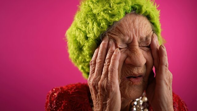 Fisheye funny view of sick tired headache migraine exhausted elderly mature old woman rubbing temples isolated on pink background in studio.