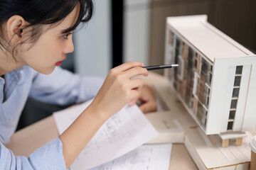 Asian architect engineer women working to examining design of architecture building model and taking notes in paper while planning construction and architectural project plan on blueprint in office