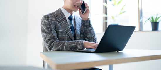オフィスでパソコン作業をしながら電話をするジャケットを着た若い男性