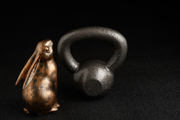Holiday fitness and Happy Easter, gold metal bunny with a silver iron kettlebell on a black background looking up adoringly
