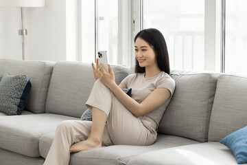 Happy beautiful young Asian blogger woman recording self video, talking selfie, talking on conference call on smartphone, using mobile phone on home sofa, holding gadget with knee support