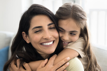 Cheerful cute daughter kid embracing happy mom from behind. Beautiful Latin mother piggybacking child with closed eyes and toothy smile, enjoying good close family relationship, motherhood