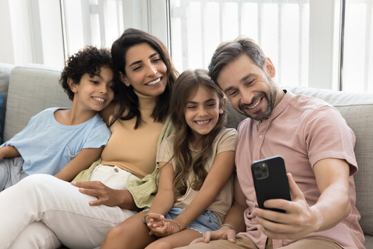 Cheerful parents and cute happy kids taking selfie, recording self video on mobile phone, having fun on home sofa, laughing, posing for frontal camera on smartphone, laughing