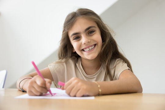 Cheerful cute early schoolgirl kid drawing pink sketches in colorful pencils, looking at camera with toothy smile, working on study school homework task, enjoying creativity, training artistic skills