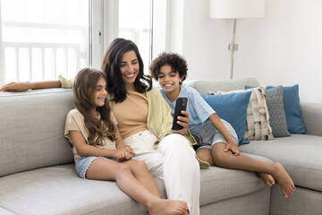 Happy mom and two sibling kids using application on mobile phone for online communication, relaxing together on couch, looking at screen, smiling, laughing, having fun, talking on video call
