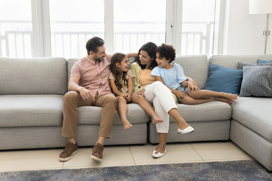 Cheerful Latin couple of parents and son and daughter kids resting on spacious comfortable couch in cozy apartment, hugging with love, joy, talking, laughing, enjoying family leisure. Wide shot